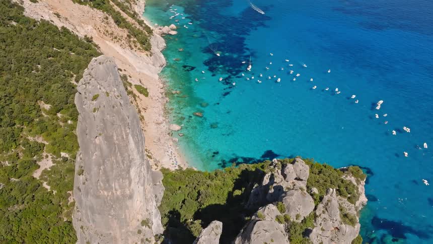 Ascending aerial footage reveals the stunning beauty of Cala Goloritzé in Sardinia, featuring its iconic rock spire, turquoise sea, and a bay full of boats