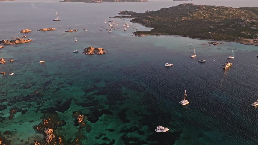 Stunning aerial footage captures a fleet of yachts and sailboats resting in the tranquil, turquoise waters of a rocky cove at golden hour