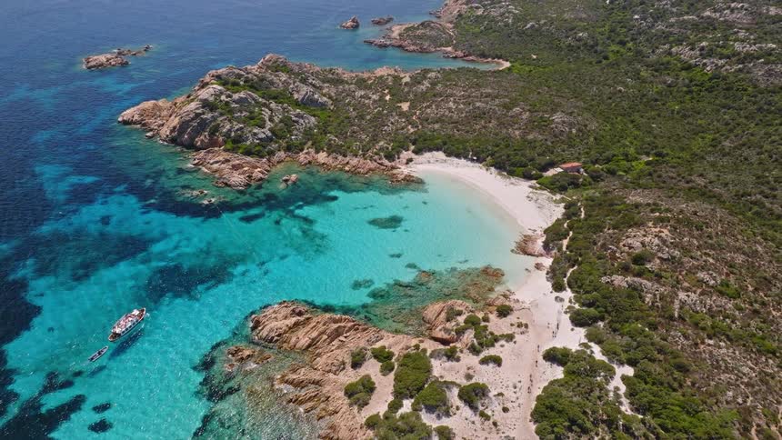 An aerial view pulls back to reveal a breathtaking secluded cove with crystal-clear turquoise water and a white sand beach on a rocky Mediterranean island