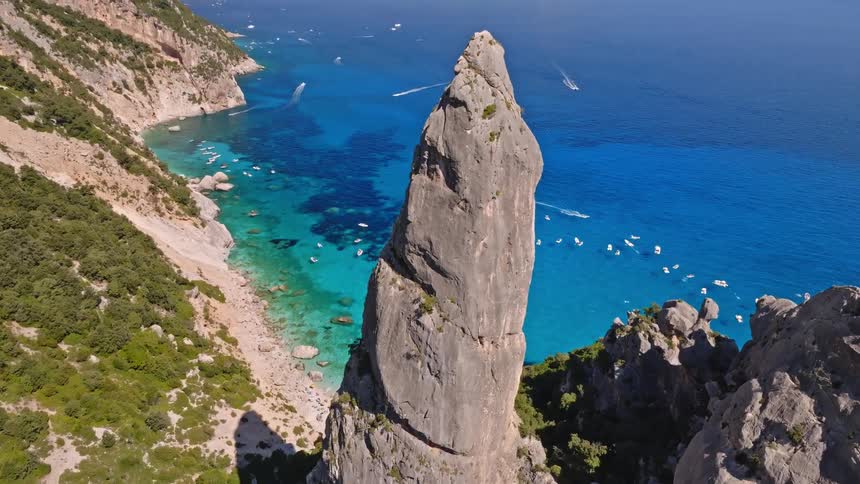 A stunning aerial view of a majestic rock pinnacle on a rugged coastline, overlooking a beautiful bay with turquoise water and many anchored boats
