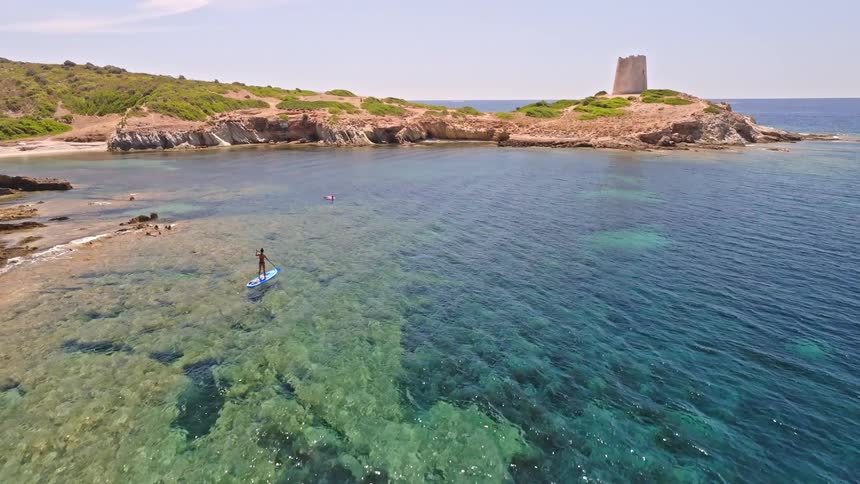 A person enjoys stand-up paddleboarding in a stunning turquoise bay with a historic stone tower on a sunny summer day
