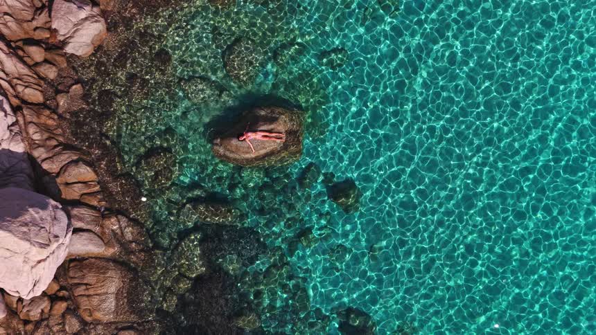 Aerial top-down view of a woman in a pink bikini sunbathing on a rock in the middle of the beautiful, clear turquoise sea