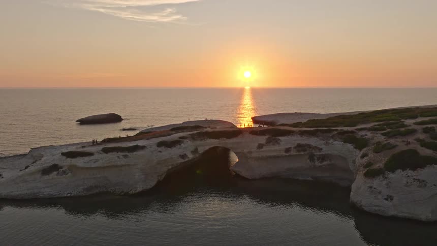 An aerial shot pulls back to reveal the sun setting through a stunning natural rock arch over the ocean
