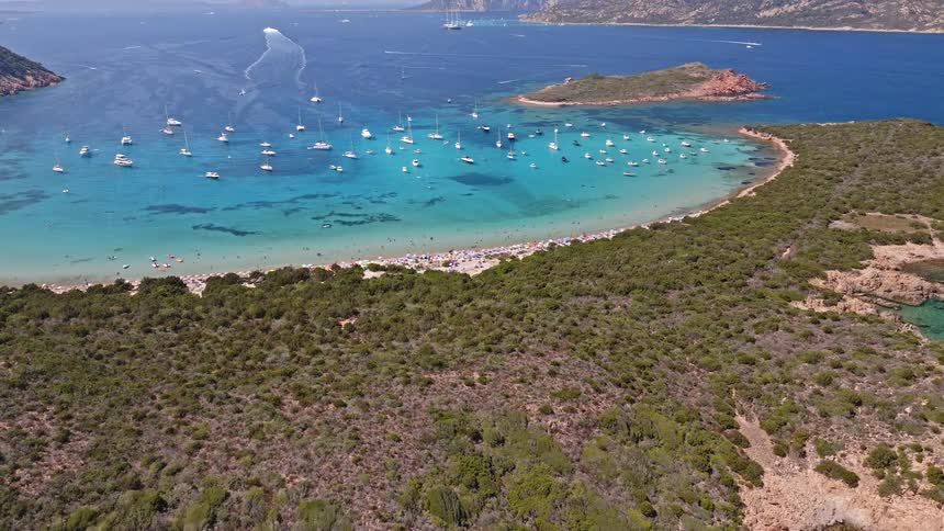 Stunning aerial view reveals a lush peninsula with a crowded beach and turquoise bay filled with sailboats on a sunny day