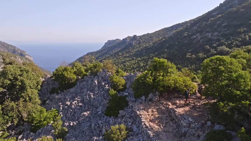An aerial view follows a couple hiking along a rugged mountain trail in a lush green valley that opens up to a stunning ocean view