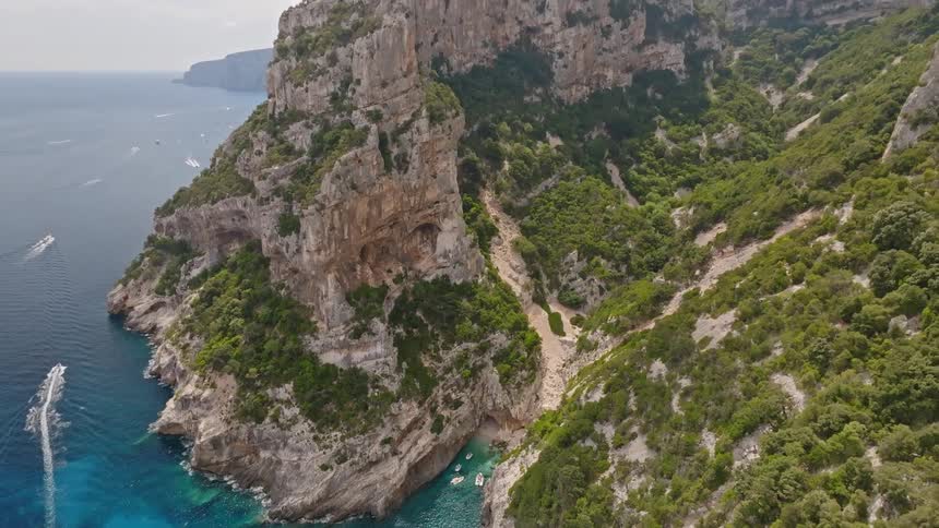 An epic aerial ascent reveals a hidden cove with boats floating on pristine turquoise water, nestled between dramatic, tree-covered cliffs on a stunning coastline