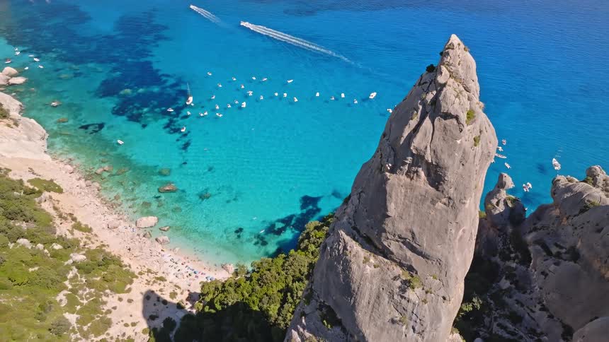 A stunning aerial view captures a towering rock pinnacle overlooking the crystal-clear turquoise bay filled with boats and a secluded beach