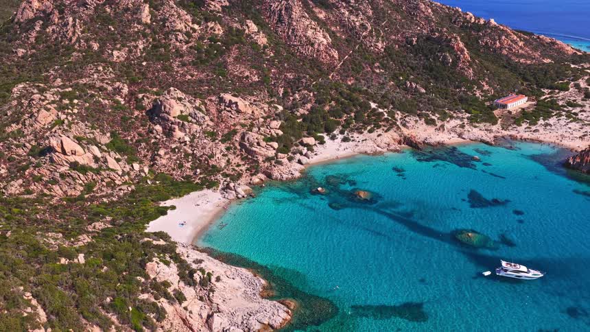 A stunning aerial view pulls back to reveal a hidden cove with crystal-clear turquoise water, a white sand beach, and a yacht anchored offshore a rocky island