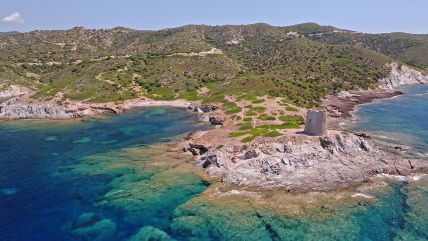 A scenic aerial shot circles an ancient stone watchtower on a rocky outcrop surrounded by the crystal-clear turquoise sea on a sunny day