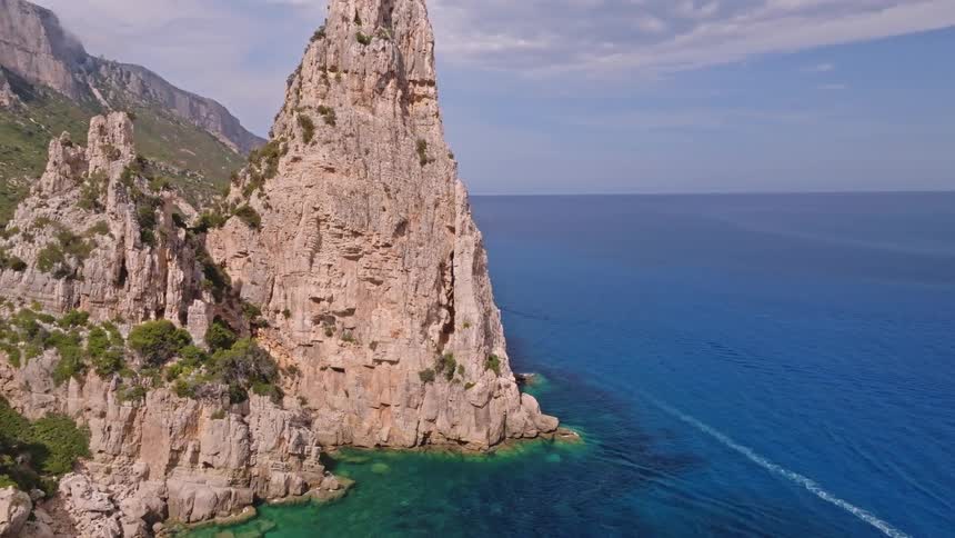 An aerial shot pulls back to reveal a majestic sea stack and the stunning turquoise coastline of Sardinia, Italy