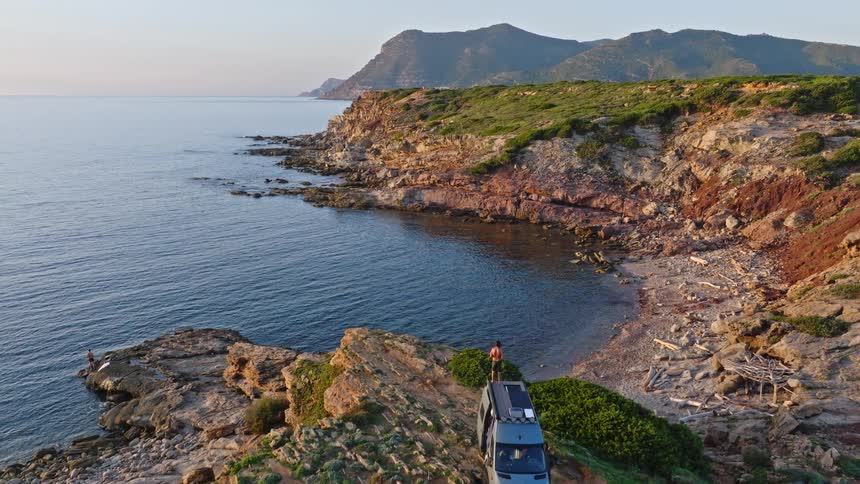 An aerial view shows a person standing on top of a campervan parked on a rugged cliff, enjoying the scenic ocean view of a secluded cove at sunrise