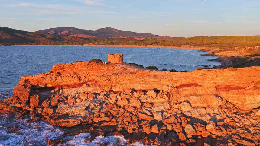 An aerial view flies towards an ancient stone tower on a rugged, red-rock coastline glowing in the warm light of a golden hour sunset