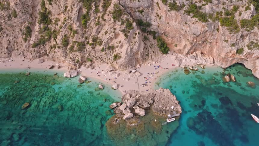 An aerial top-down view slowly pulls back from a stunning secluded beach with turquoise water nestled at the base of a massive rocky cliff