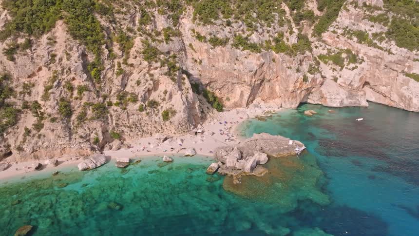 A stunning high-angle aerial view slowly reveals a secluded beach cove with turquoise waters nestled beneath towering cliffs