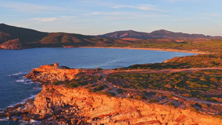 Golden hour light illuminates the stunning red cliffs and historic watchtower overlooking a tranquil bay in this beautiful aerial shot