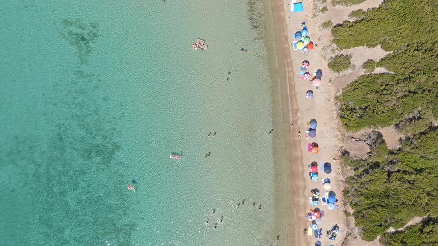 An aerial top-down perspective captures tourists swimming in the pristine turquoise sea and sunbathing under colorful umbrellas along a sandy shoreline