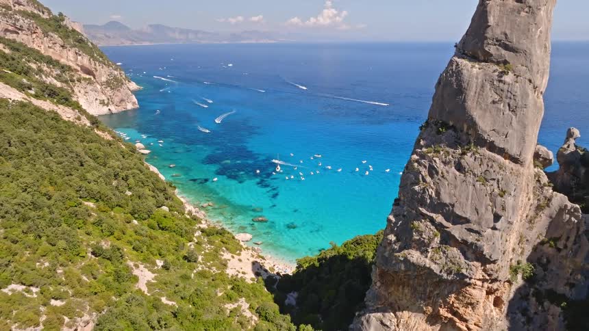 An aerial pullback shot reveals the iconic Aguglia pinnacle towering over the stunning turquoise waters of Cala Goloritzé beach in Sardinia, Italy