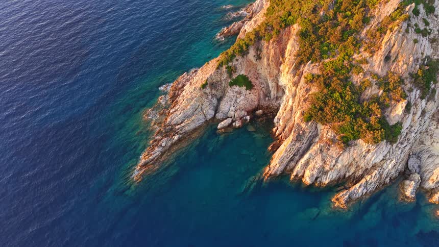 An aerial pullback reveals a majestic, rocky island coastline bathed in golden sunrise light over the calm, deep blue sea