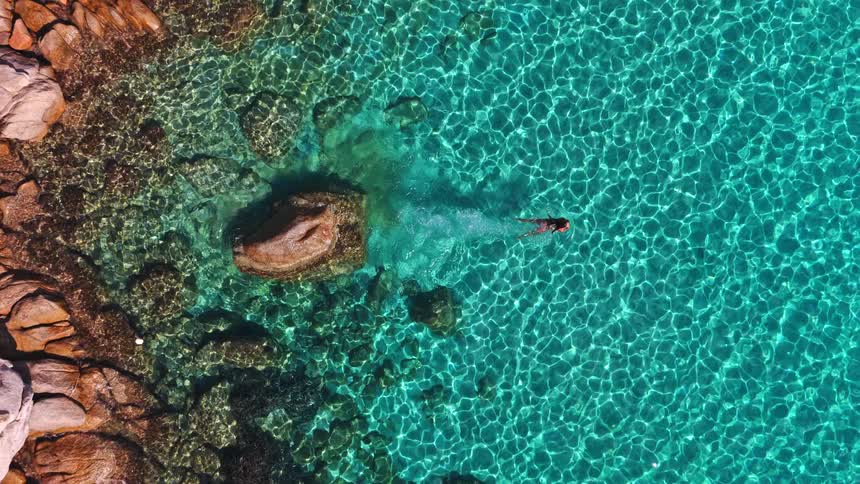 An aerial top-down view captures a woman as she gracefully dives from a sun-drenched rock into the shimmering, crystal-clear turquoise sea and swims along the coastline