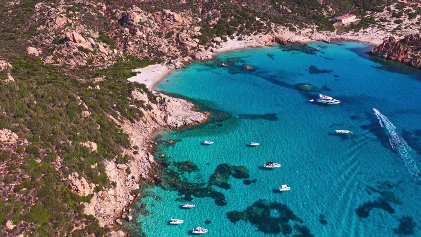 An aerial view reveals a stunning secluded cove with crystal-clear turquoise water where yachts are anchored near a rocky island