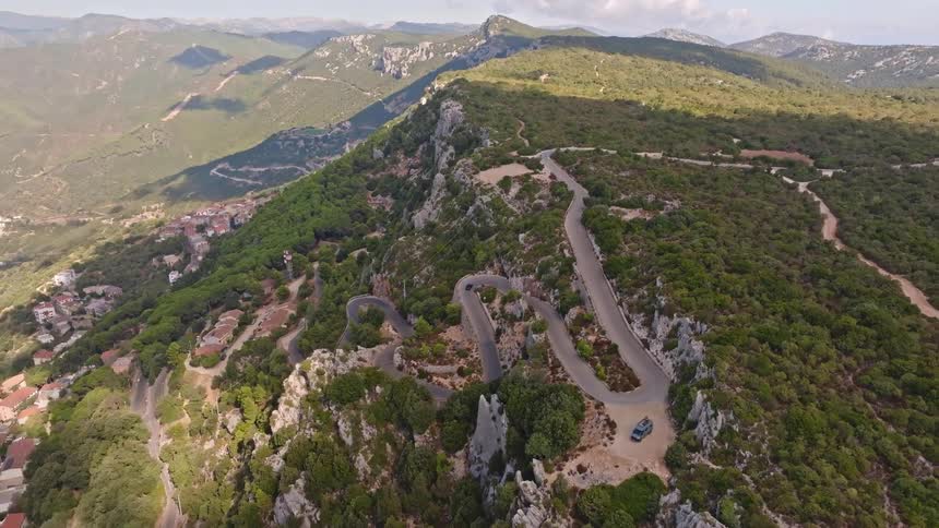 An aerial drone pulls back to reveal a winding serpentine road on a rugged, tree-covered mountain under a partly cloudy sky