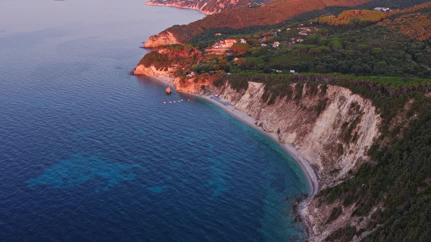A stunning aerial view tilts up to reveal a beautiful and rugged island coastline with sheer cliffs and a secluded beach, all bathed in the warm light of sunset