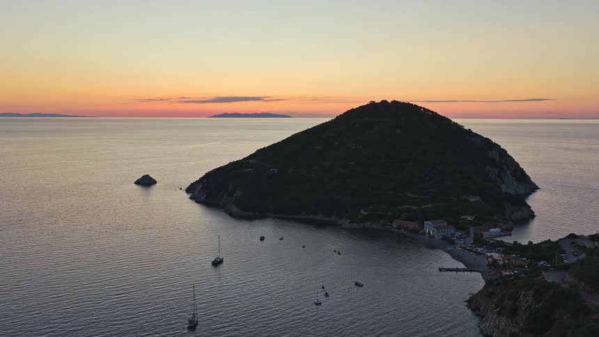 Stunning aerial pull-back reveals a beautiful coastal island and a small village nestled in a calm bay at sunset