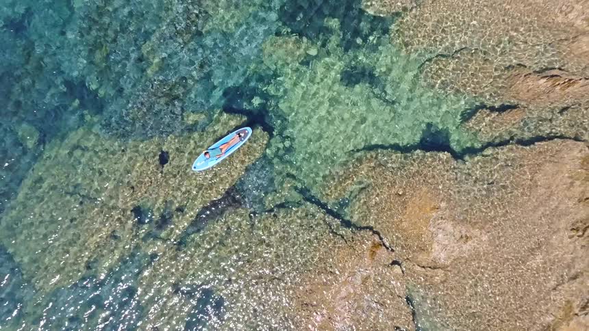 A tranquil aerial top-down view shows a woman peacefully relaxing on a paddleboard in the crystal clear turquoise ocean, as the drone slowly ascends