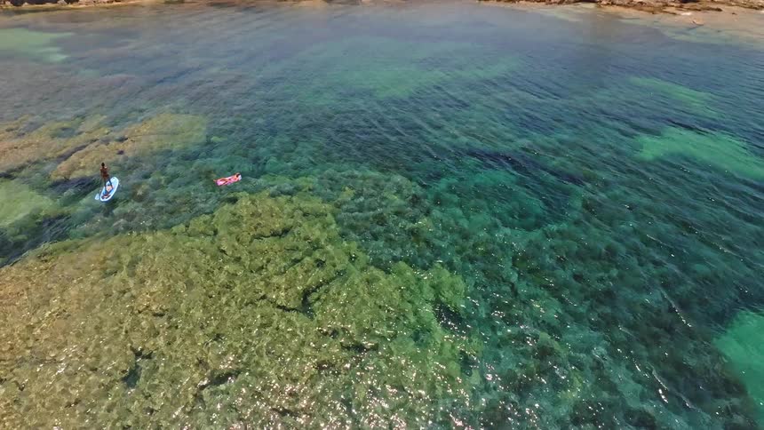 An aerial view tilts up to reveal people paddleboarding and floating in the clear turquoise sea next to a rocky island with a historic tower