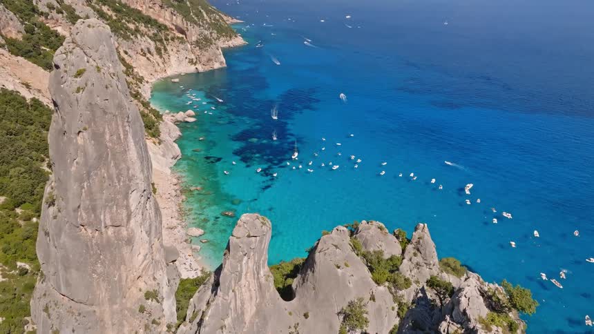A breathtaking aerial view rises over dramatic rock spires and cliffs, revealing a stunning turquoise sea filled with yachts along a sunny Mediterranean coastline