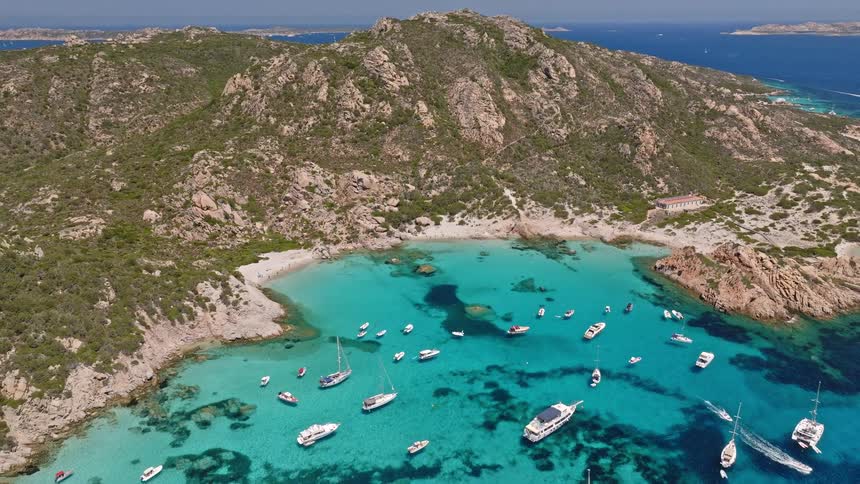 Stunning aerial view flying away from a beautiful secluded beach and turquoise cove filled with anchored boats and yachts on a sunny day