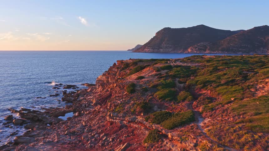 An aerial view flies over a rugged, rocky coastline illuminated by the warm golden light of sunrise, with the deep blue ocean stretching to the horizon