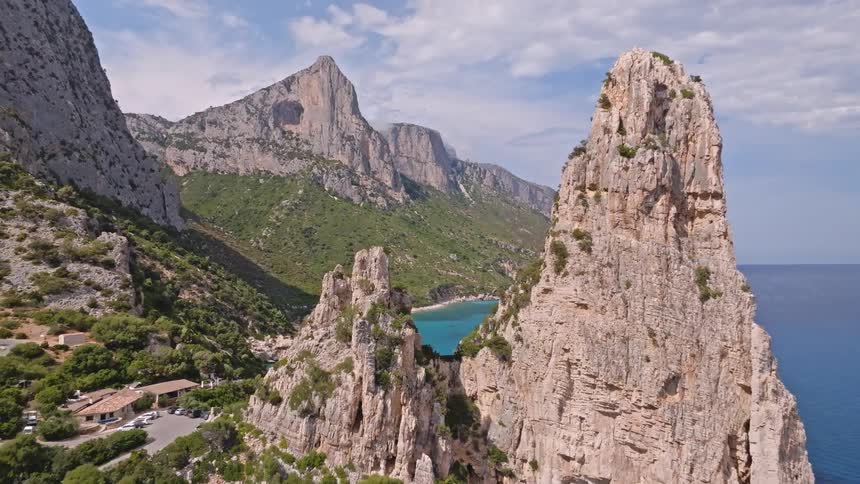 An aerial view pulls back from a turquoise bay to reveal the dramatic, rocky coastline and a prominent spire of Aguglia di Goloritze in Sardinia, Italy