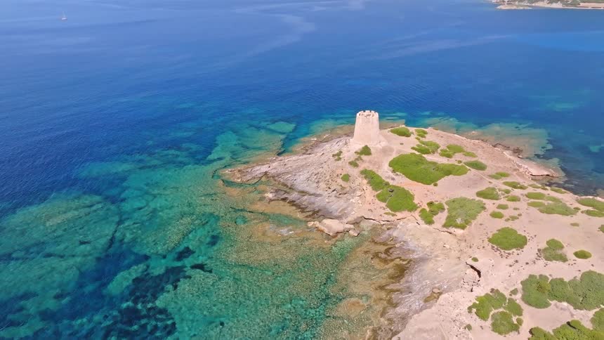 Stunning aerial pullback shot reveals a historic stone tower on a remote rocky peninsula surrounded by crystal-clear turquoise waters