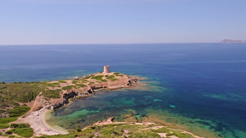 Stunning aerial footage rises over a verdant coastline to reveal an ancient stone tower on a peninsula with crystal-clear turquoise waters