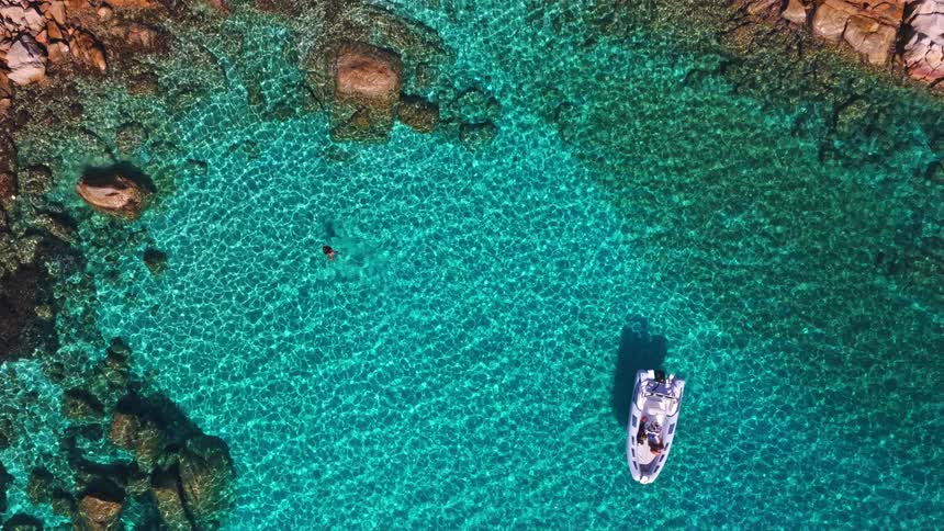 Aerial top-down view reveals a swimmer and a boat in a stunningly clear turquoise sea cove on a sunny day