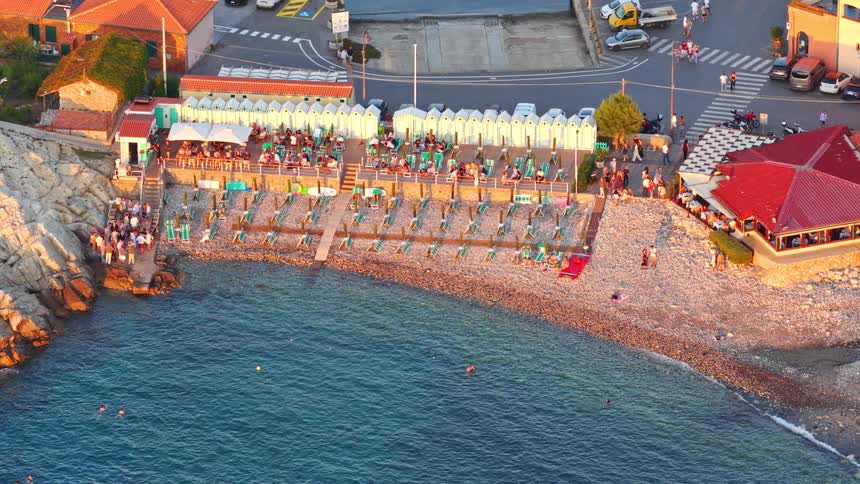 A static high-angle view of a bustling Italian beach club on a rocky coast during a beautiful golden hour sunset