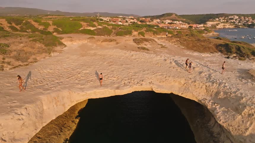 Descending aerial shot reveals a group of people standing on a beautiful natural rock arch over the sea during a golden sunset