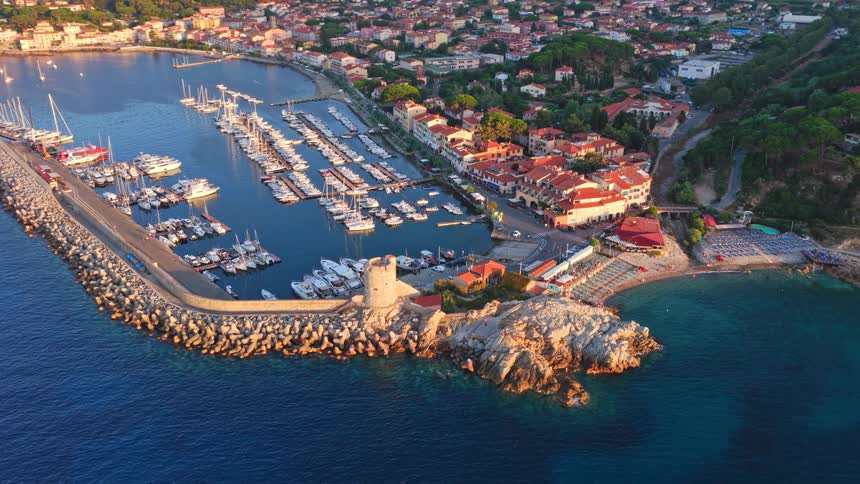 A stunning aerial view reveals the golden light of sunset illuminating the charming harbor, historic tower, and coastal town of Marciana Marina, Elba