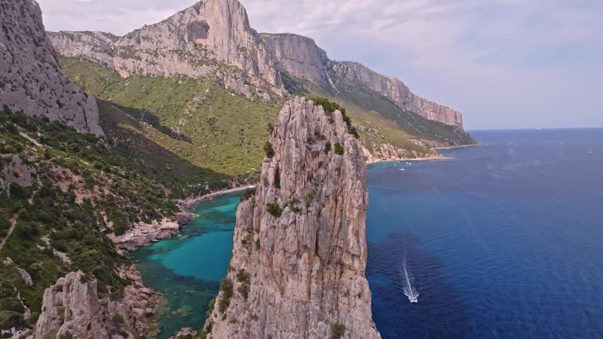 A breathtaking reverse aerial shot reveals the iconic Aguglia Goloritze sea stack on the stunning, rugged coastline of Sardinia, Italy