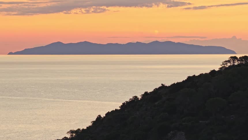 Stunning aerial footage pans over a tranquil sea at sunrise, revealing a silhouetted island against a warm orange sky