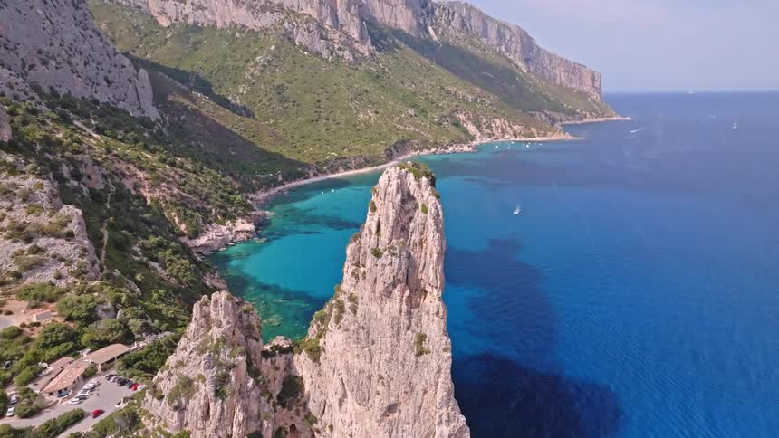 A top-down aerial view tilts up to reveal the magnificent limestone pinnacle of Aguglia di Goloritzè on the rugged Sardinian coastline, surrounded by a crystal clear turquoise sea