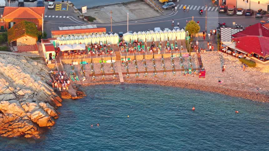 A stunning aerial view captures a bustling beach club and restaurant on a rocky coastline during a picturesque golden hour sunset