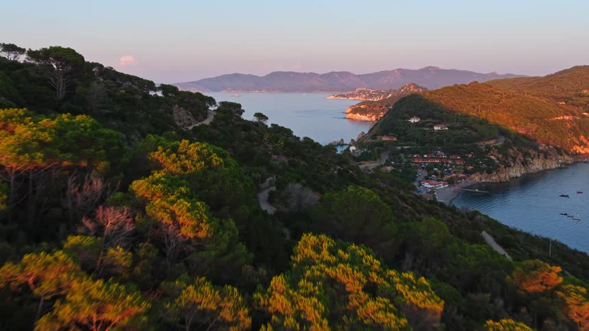 An aerial view flies over the sun-kissed, tree-covered hills of Elba Island, Italy, revealing a picturesque coastal town and bay at golden hour