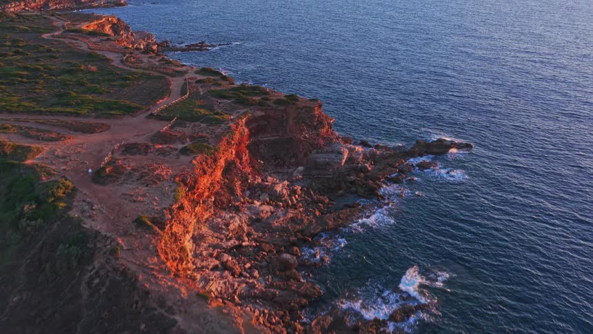 An aerial flyover reveals a historic stone tower on a rugged, sun-drenched coastline at golden hour