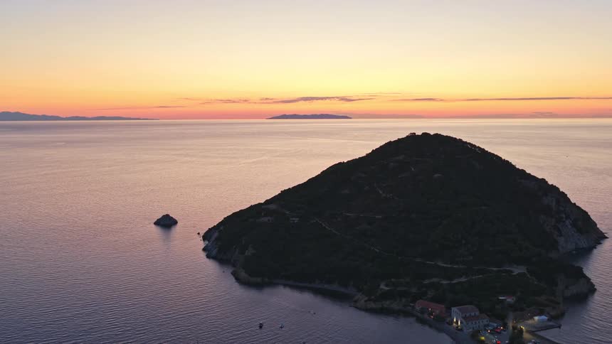 An epic aerial pullback shot captures the beautiful colors of sunset over the sea, revealing a tranquil island village