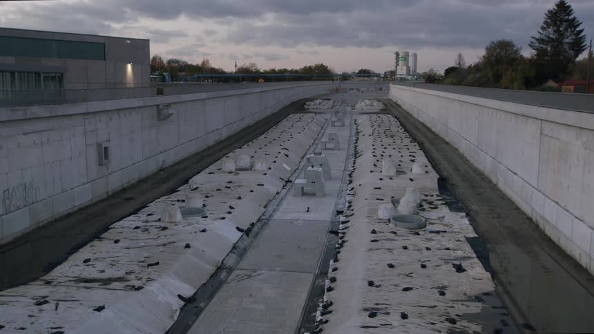 A wide, static shot shows a massive, empty construction site for a road or railway within a concrete underpass at dusk