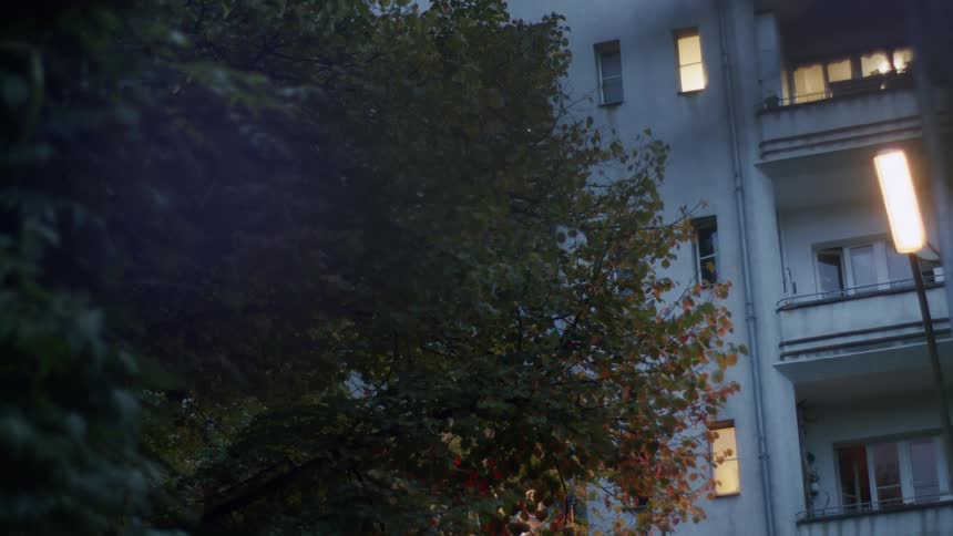 A quiet exterior view of an apartment building at dusk with windows lit and a street lamp glowing warmly beside the trees