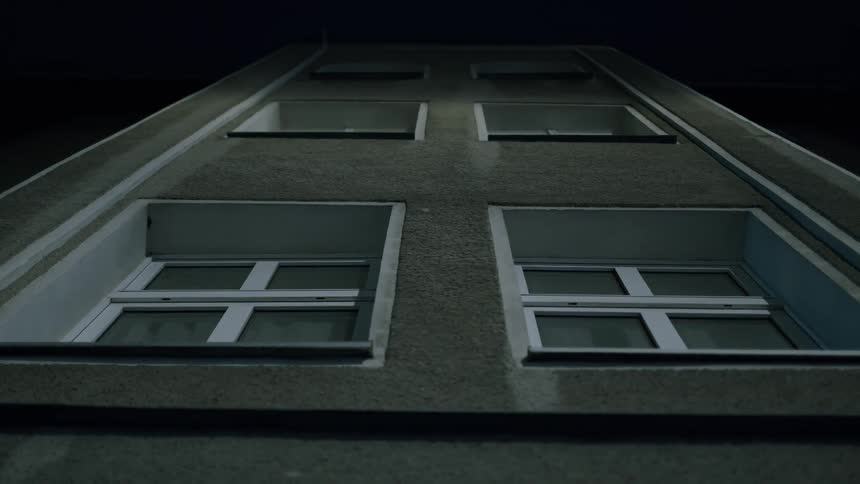 A static, low-angle shot looks up at the windows of a stark building facade against the dark night sky