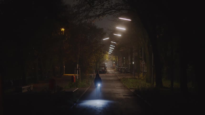 A lone cyclist rides toward the camera with a bright headlight on a dark, foggy, tree-lined path at night